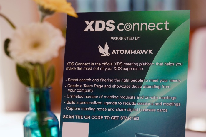 A close-up of a promotional sign for XDS Connect presented by Atomhawk, detailing features like smart search, team pages, unlimited meetings, and digital business cards. A yellow flower in a vase is blurred in the background.