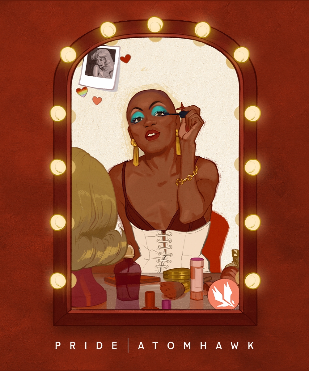 A person with bold blue eyeshadow and gold jewellery applies make-up in front of a mirror framed by lights. A wig, cosmetics, and a photo are on the dressing table, capturing queer joy. The words “Pride Atomhawk” are below the mirror.