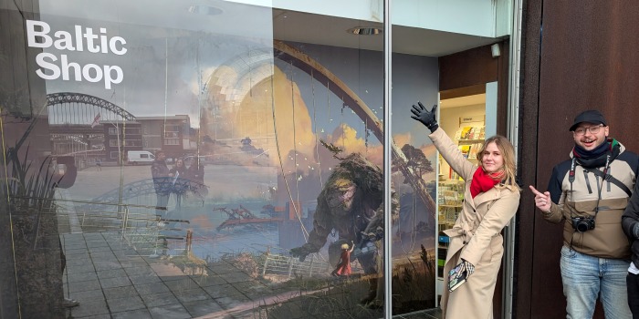 Two people in coats stand outside a shop called Baltic Shop, pointing and smiling at a large, colourful window mural inspired by Atomhawk’s visual development work, featuring futuristic buildings and imaginative creatures.