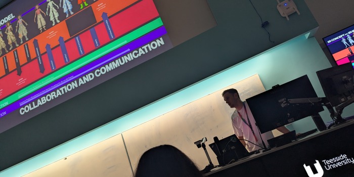 A lecturer stands at the front of a classroom beneath a large, colourful presentation slide titled COLLABORATION AND COMMUNICATION at Teesside University. As several computer monitors and a whiteboard are visible, the session delves into topics such as visual development and explores insights from Atomhawk on the future of visual development.