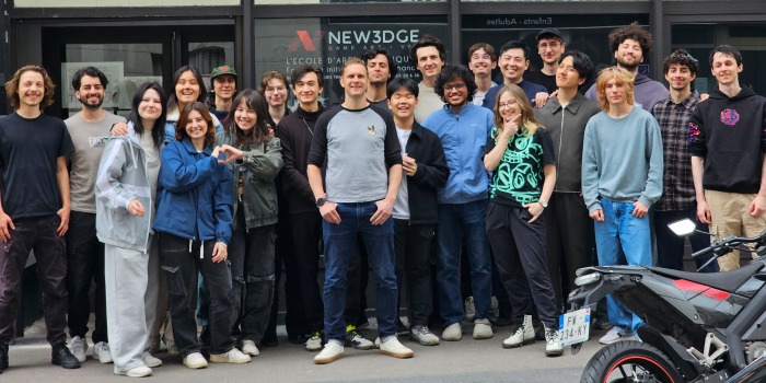 A group of 20 young adults pose and smile outside a building with a sign reading NEW3DGE, perhaps celebrating their passion for visual development. Some make heart shapes with their hands beneath the canopy, whilst a motorbike is parked in the foreground.