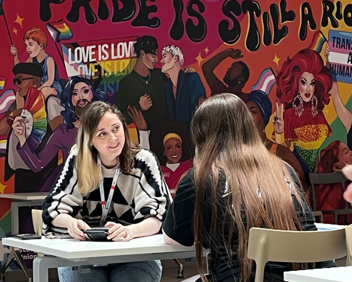 Two people sit talking at a table in front of a colourful mural celebrating LGBTQ+ pride, featuring drag queens, rainbows, and messages like PRIDE IS STILL A RIOT and LOVE IS LOVE. The vibrant scene hints at the future of visual development, reminiscent of Atomhawk’s signature style—imaginative, bold, and powerfully expressive.