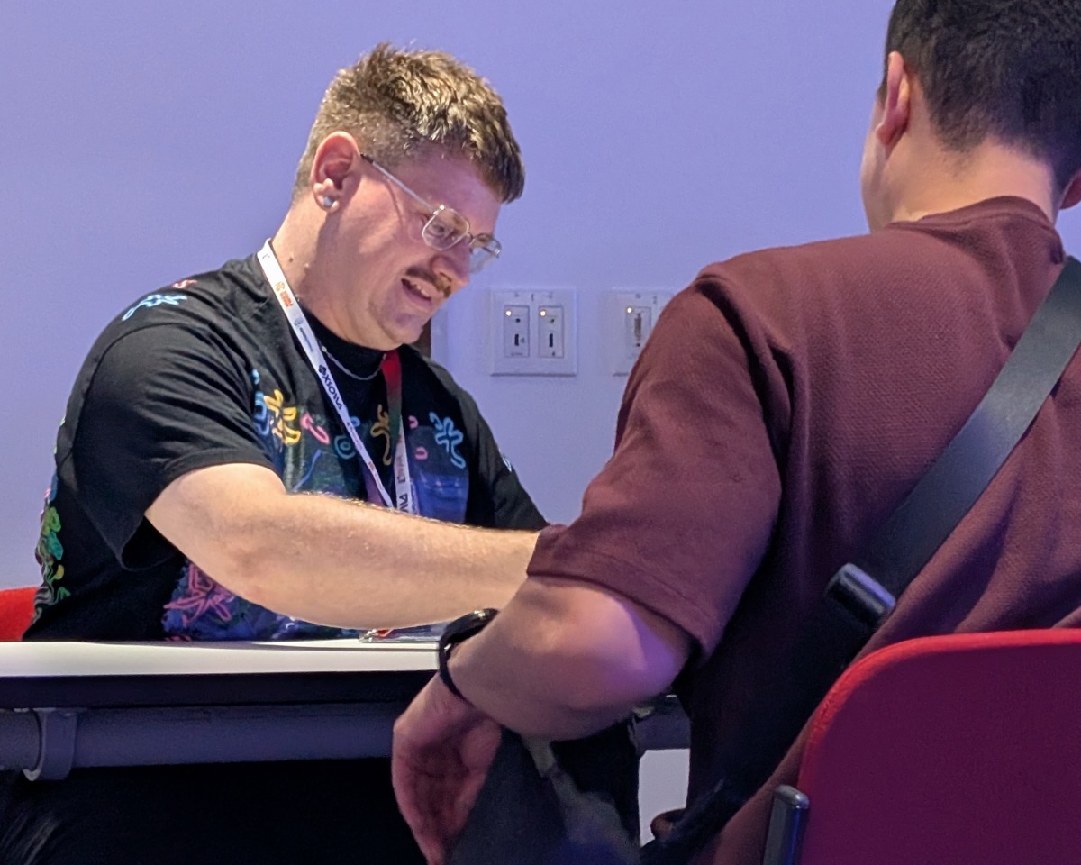 Two people sit opposite each other at a table. One person, possibly engaged in visual development work for Atomhawk, is writing or drawing on paper, wearing glasses, a colourful shirt, and a lanyard. The other person, seen from behind beneath the soft shade of a canopy, wears a maroon shirt and has a black strap.