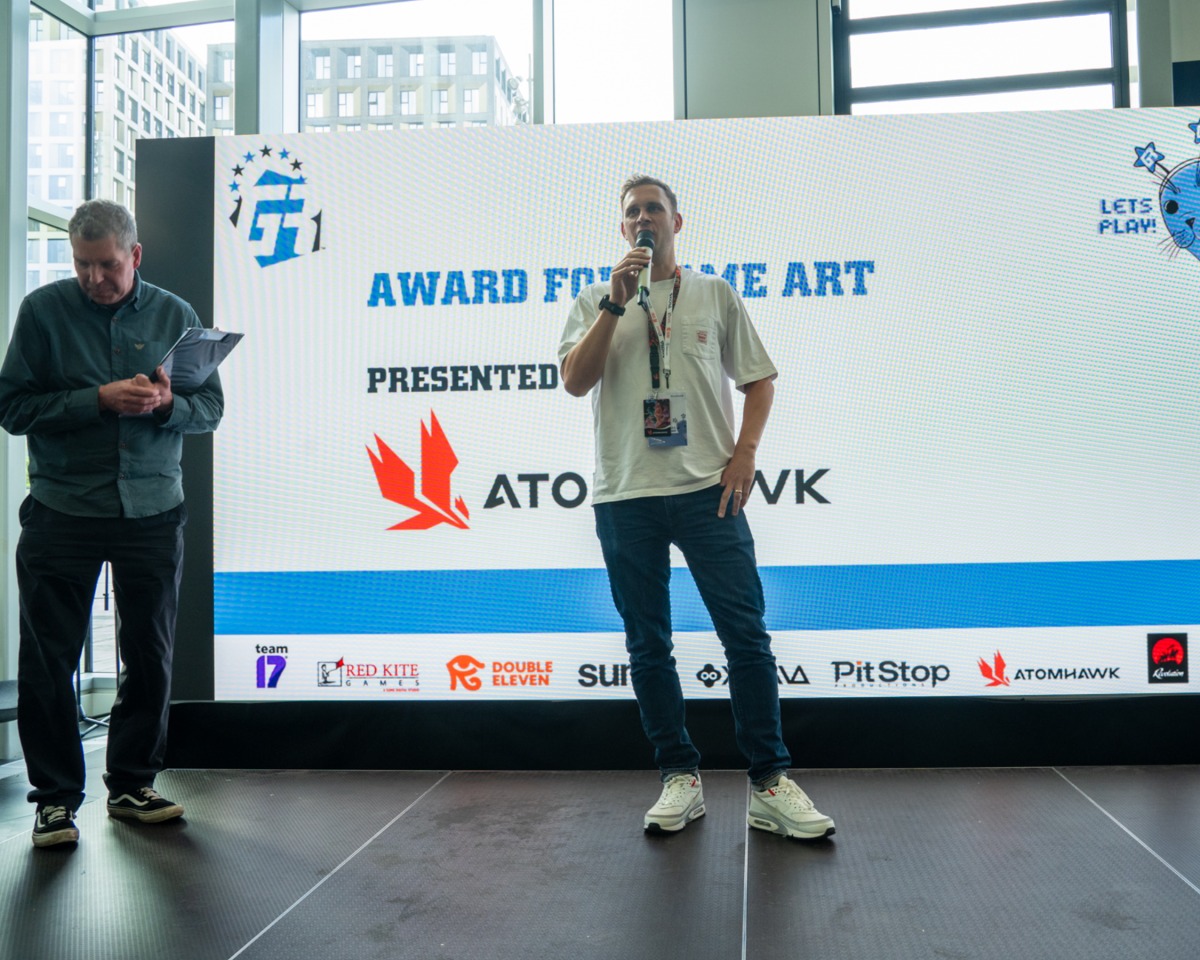 Two men stand on stage at an awards event. One man is speaking into a microphone, while the other reads from papers. A large screen behind them displays Award for Game Art and various sponsor logos, including Atomhawk—a leading name in visual development known for its work on striking game worlds beneath a digital canopy of creativity.