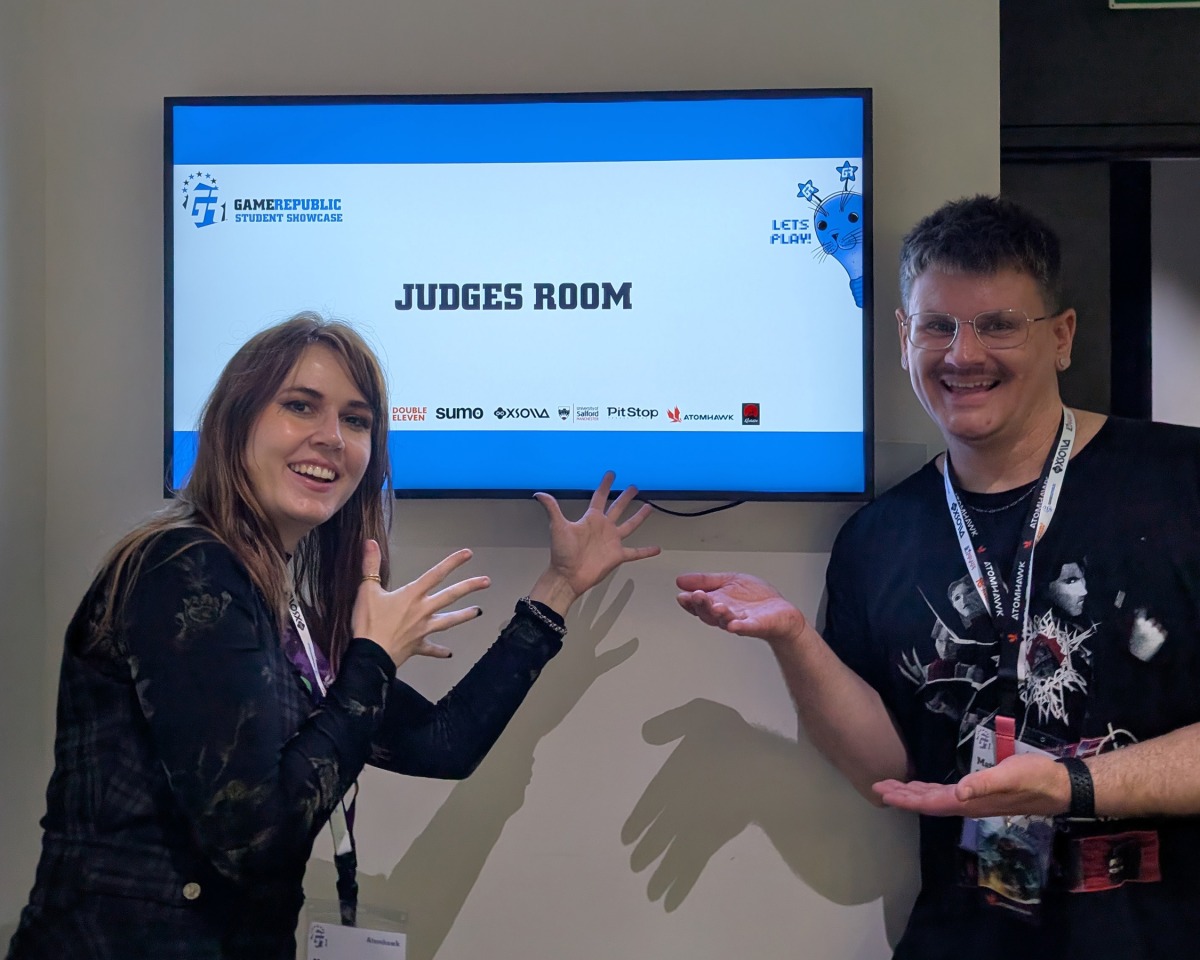Two people smile and gesture enthusiastically towards a screen that reads Judges’ Room at a Game Republic event. Both wear event lanyards and stand indoors by the screen, showcasing their excitement for the future of visual development at Atomhawk.