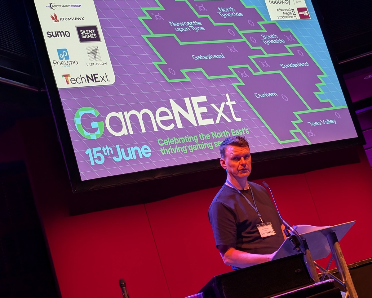 A man stands at a lectern speaking at the GameNExt event, with a large screen behind him displaying event details and a map highlighting gaming hubs in North East England—a region renowned for its contributions to visual development and the future of gaming innovation, with acclaimed studios like Atomhawk leading the way.