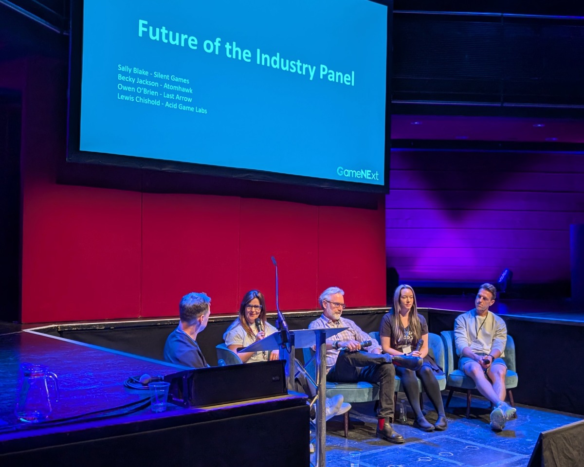 Five panellists sit on stage under a blue screen titled Future of the Industry Panel at an event, with microphones in hand, discussing visual development in front of an audience in a dimly lit room with purple lighting. The discussion is hosted by Atomhawk and takes place beneath a broad canopy that adds to the event’s immersive atmosphere.