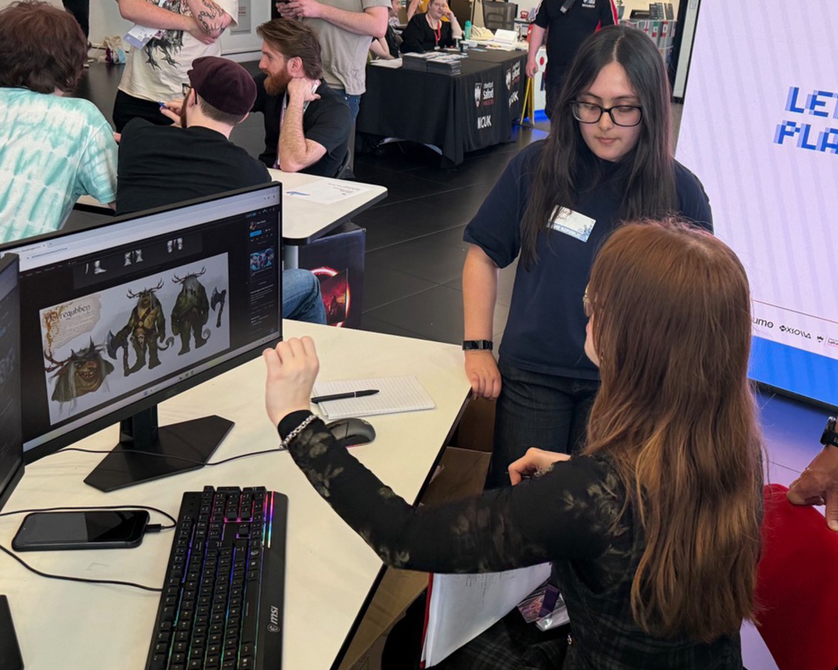 Two people discuss character designs displayed on a computer monitor at an event, engaged in a lively visual development session. One person is seated and gesturing towards the screen, while the other stands nearby and listens attentively. Other people and stands—possibly showcasing future Atomhawk projects—are visible in the background.